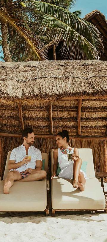 A man and woman sitting on cabanas under palm trees on the beach