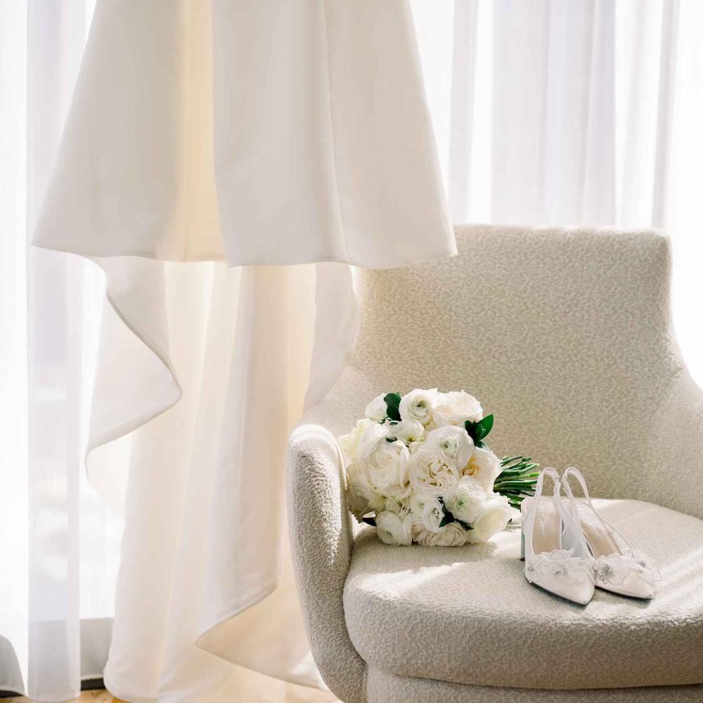 A veil hung up near a white armchair with a bridal bouquet and shoes resting upon it