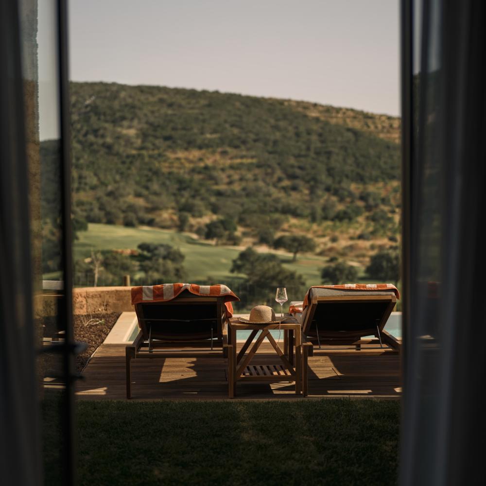 A view of two poolside recliners through a patio window, with the Algarve hills in the background