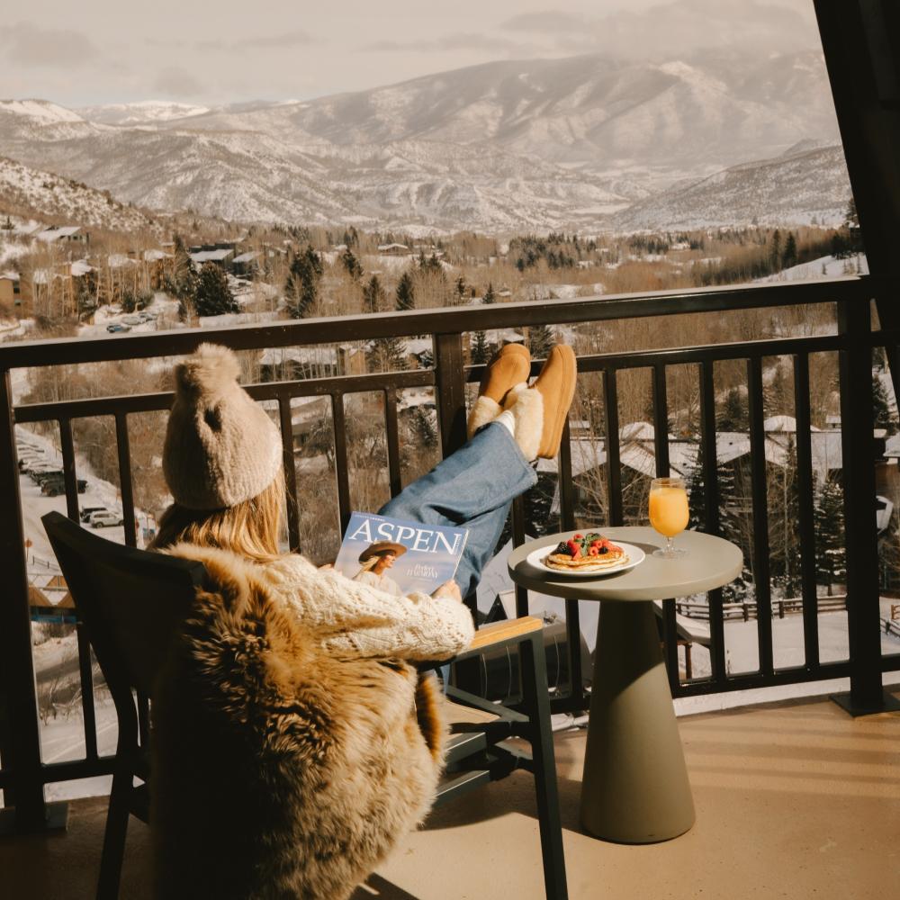 A woman reads a magazine seated in a chair on a balcony overlooking a snowy Colorado mountain range