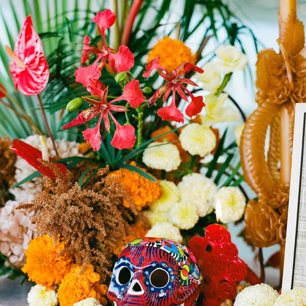 Brightly painted red sugar skull sits before a vibrant Day of the Dead altar arrangement featuring orange marigolds, red flowers, and green foliage next to a tall, molded brown candle.
