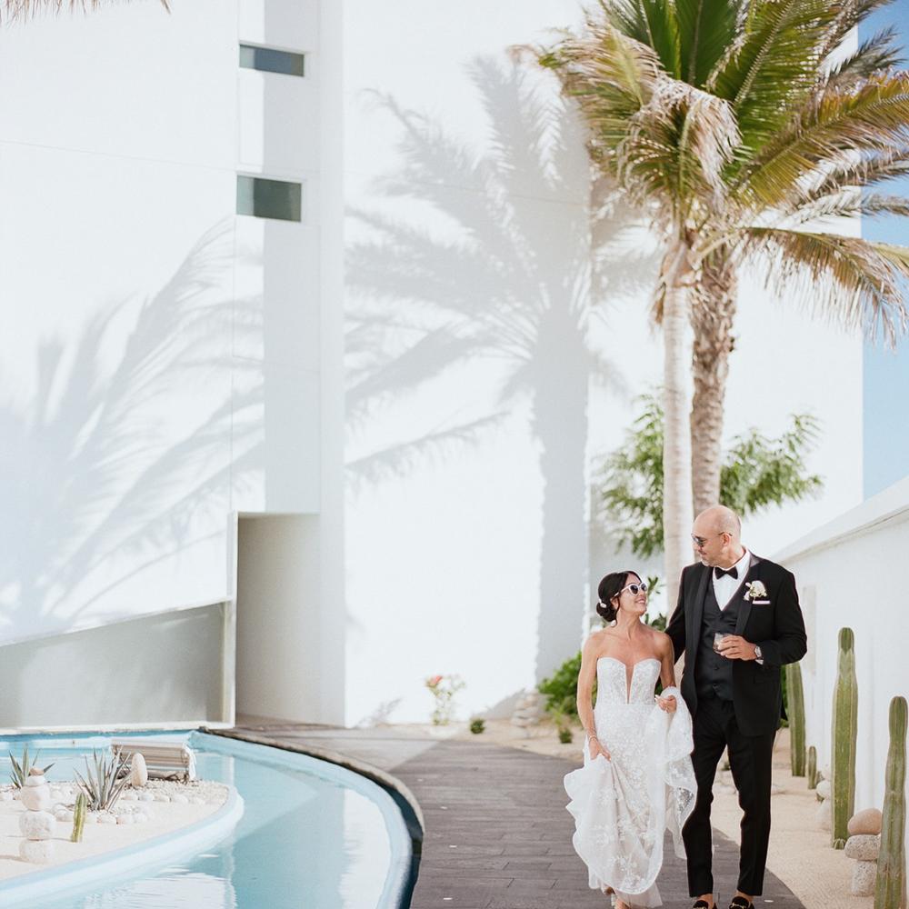 A newly married couple in wedding attire walks along a dark pathway beside a bright blue pool, framed by a white building and tall palm trees casting distinct shadows.