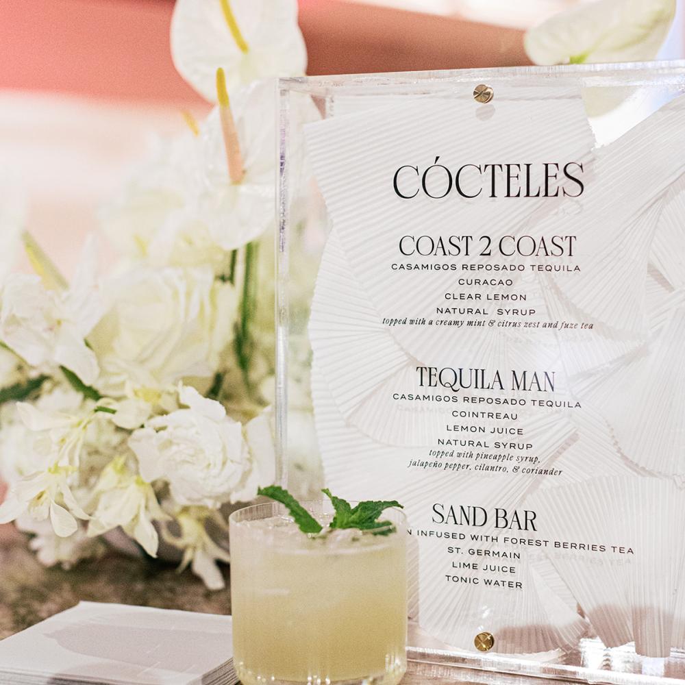 A clear acrylic cocktail menu titled "CÓCTELES" stands next to a glass of pale green iced drink garnished with mint, with white floral arrangements blurred in the background.