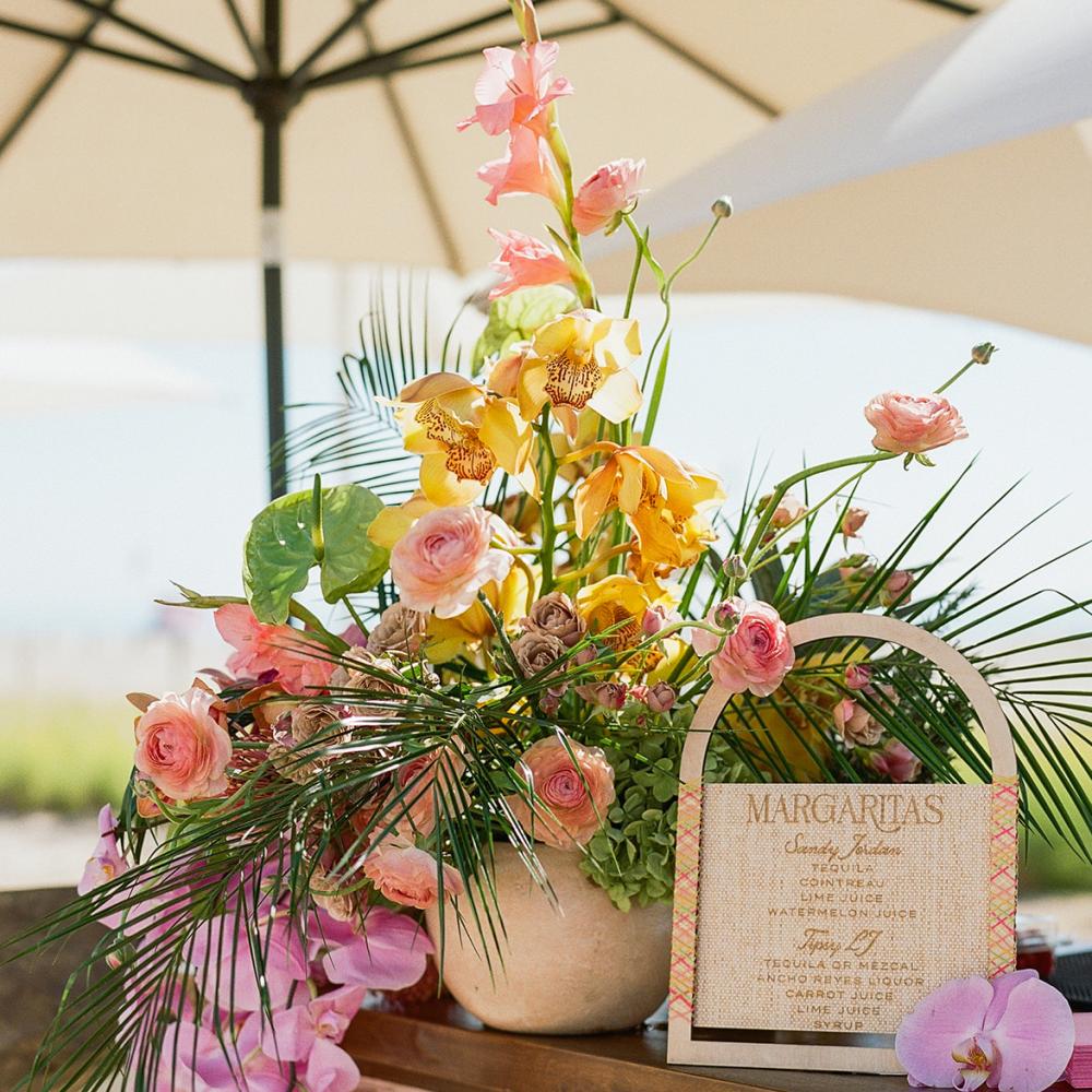 Lush floral arrangement featuring yellow orchids, pink roses, and tropical greenery next to a wooden-framed margarita menu sign on a pink tiled outdoor bar under a large umbrella.