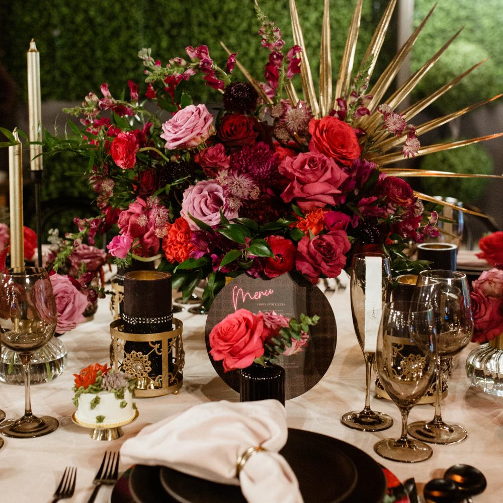 A richly decorated table setting features a dramatic centerpiece of deep red and pink roses against a green hedge backdrop, complemented by gold accents and dark tableware.