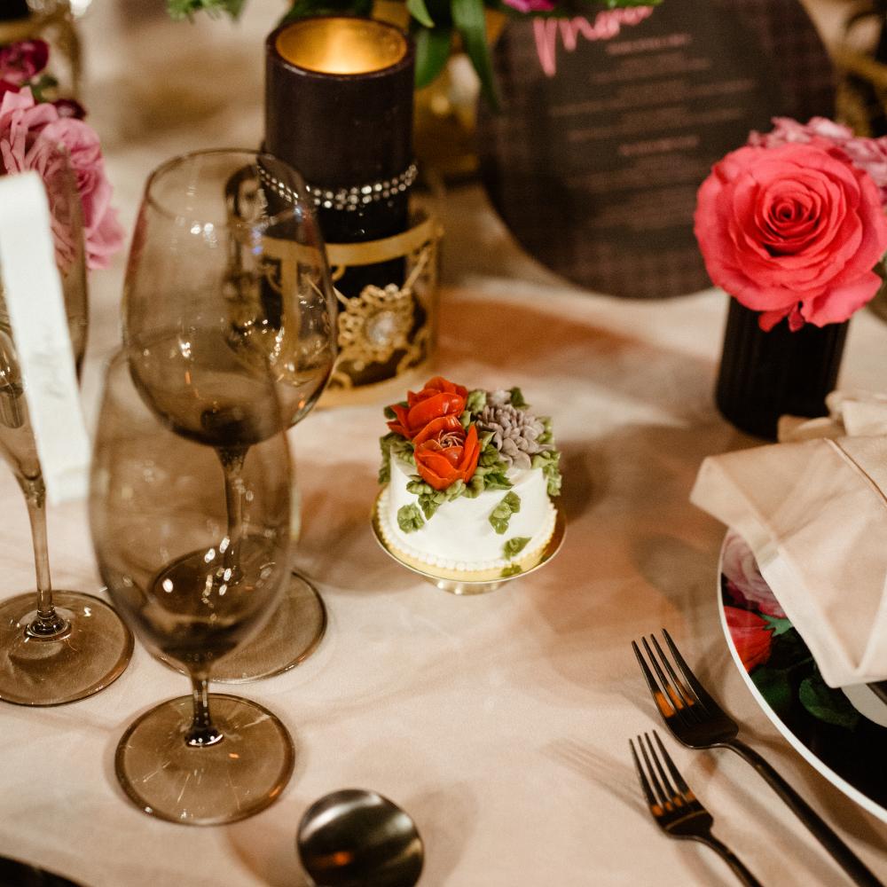 A formally set table features tinted wine glasses, black cutlery, a small white cake with red and green floral frosting, a lit dark candle, and a vibrant pink and red floral centerpiece.