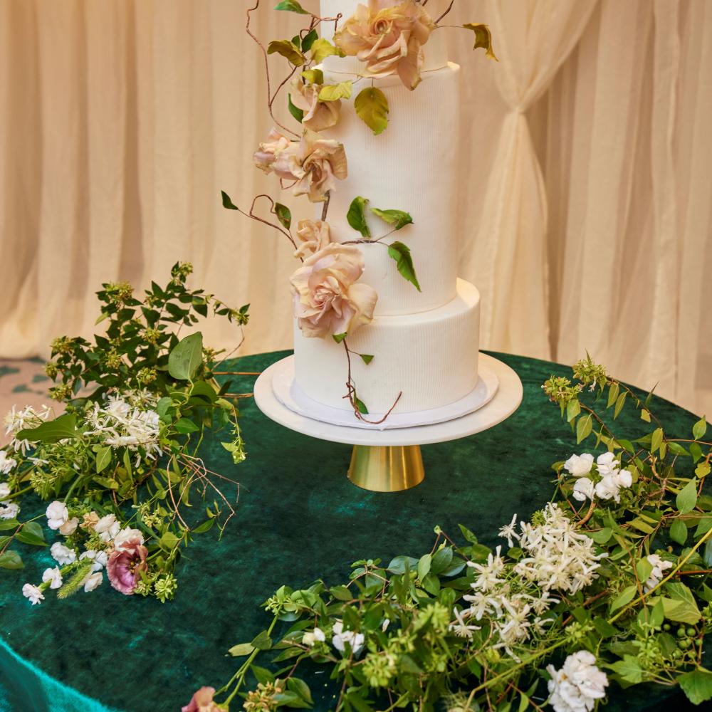 Three-tiered white wedding cake adorned with blush sugar roses and trailing vines, set on a gold stand atop a dark emerald velvet draped table surrounded by lush green and white floral arrangements.