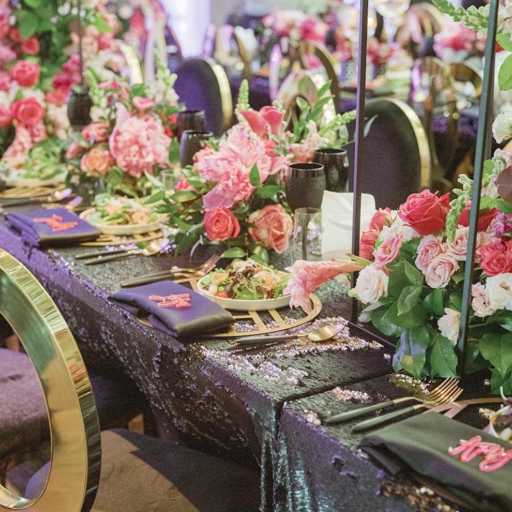 Uma mesa de banquete ricamente decorada apresenta uma toalha preta com lantejoulas, centros de mesa altos com arranjos florais em tons de rosa e vermelho, talheres dourados e guardanapos escuros com bordados coloridos.
