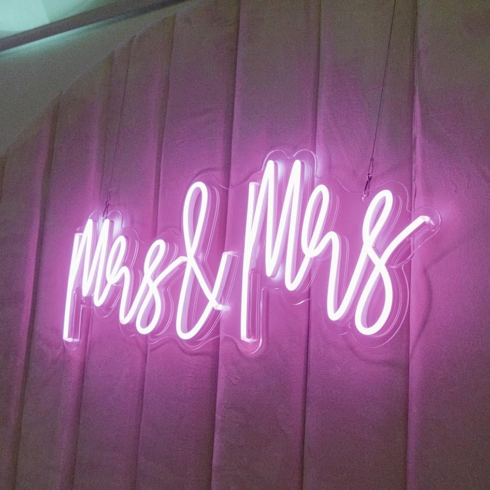 Um letreiro em neon rosa vivo com a inscrição «Mrs & Mrs» ilumina um pano de fundo drapeado, com textura e em tons de rosa-malva.