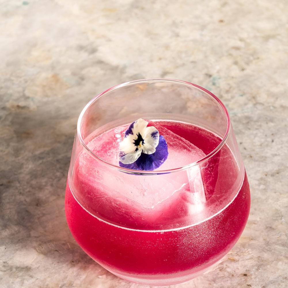 Um cocktail de cor rosa vibrante, servido num copo arredondado, repousa sobre uma superfície de mármore claro, com um grande cubo de gelo decorado com uma pequena flor comestível roxa e branca.