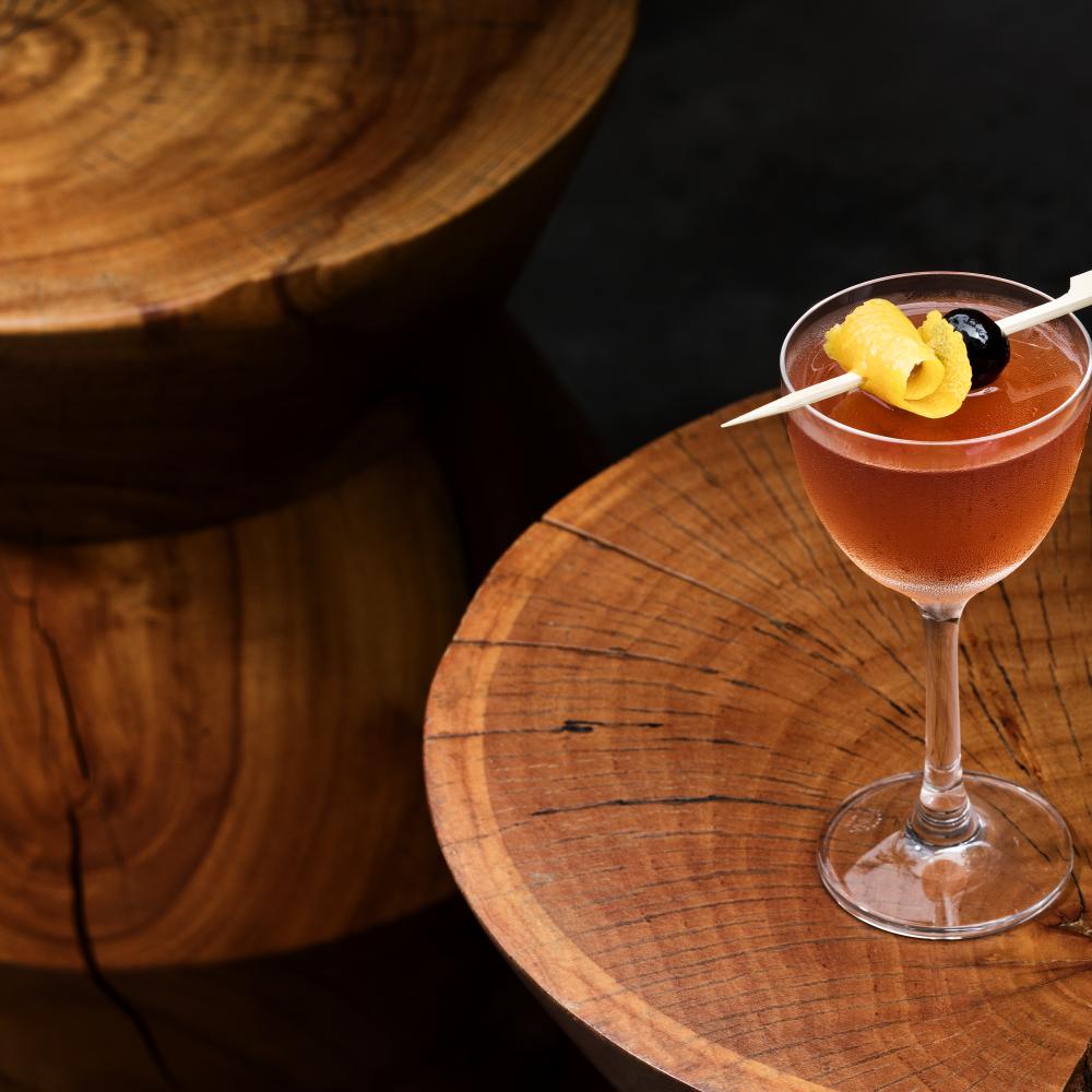 Um copo de coupé com um cocktail âmbar, decorado com uma casca de limão enrolada e uma cereja preta, repousa sobre uma mesa redonda feita de uma fatia de madeira rachada, ao lado de bancos de madeira empilhados.