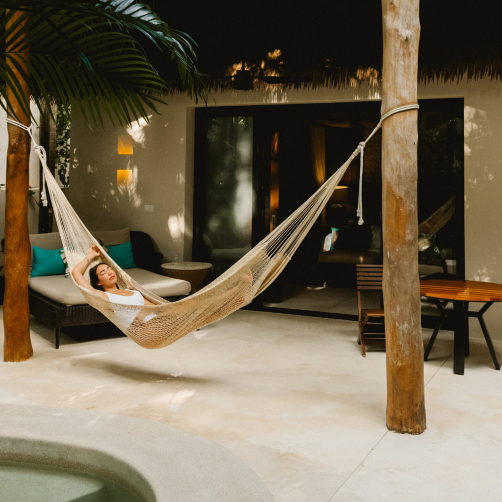Une femme se détend dans un hamac sur une terrasse extérieure agrémentée de palmiers et d'une piscine.