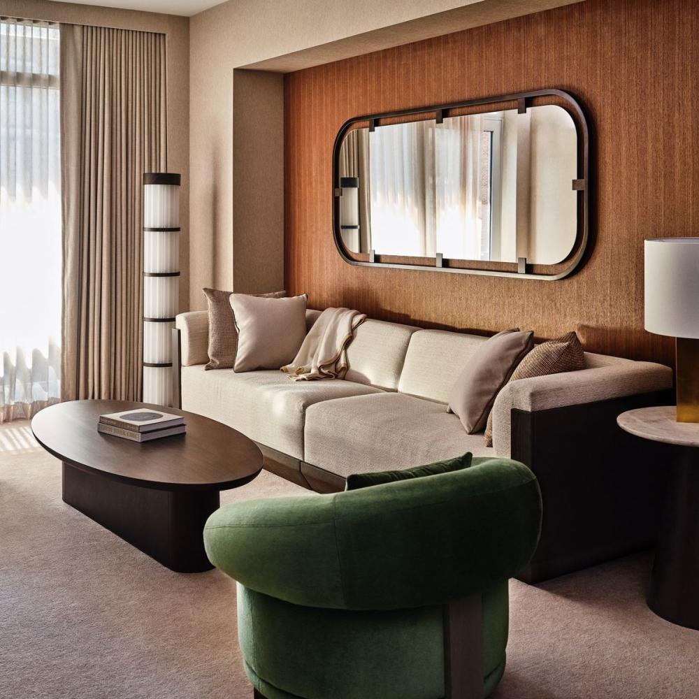 Modern living room with a cream sofa, green armchair, oval coffee table, floor lamp, wall mirror, and tall curtained windows.