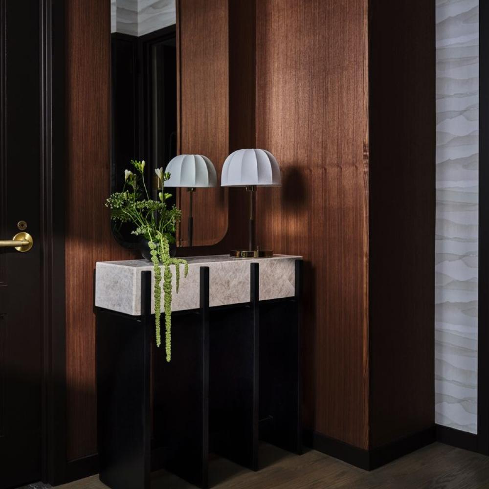 A dark wood entryway features a rounded mirror, black console table, two dome lamps, and a vase of trailing greenery.