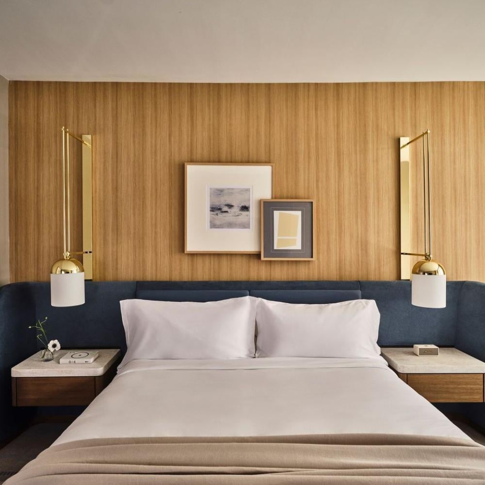 A neatly made bed with white linens sits against a curved navy upholstered headboard, flanked by floating nightstands and brass wall lamps.