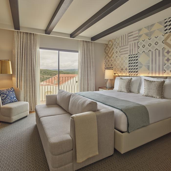 A King Bed in a large suite, with a chaise at the foot of the bed, two bedside tables with lamps, and a reading nook with an armchair and standing lamp, with light streaming in from the Algarve countryside through a sliding door 