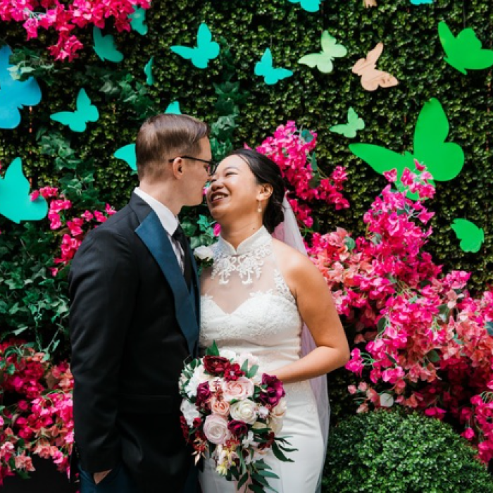 A smiling bride in a white dress and groom in a black tuxedo stand before a vibrant green wall decorated with bright pink flowers and colorful butterfly cutouts.
