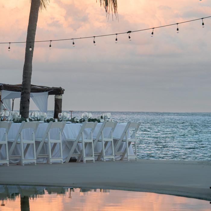Un décor de réception de mariage, composé de chaises blanches et d'une longue table, est installé sur une plage de sable, avec l'océan et des nuages aux teintes pastel en arrière-plan.