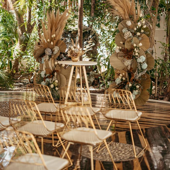 Décor de cérémonie de mariage composé de rangées de chaises dorées, de compositions florales séchées et d'une petite table, le tout dans un cadre tropical luxuriant.