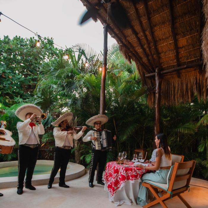 Un groupe de mariachis joue pour une femme assise à une table dans un décor tropical en plein air.