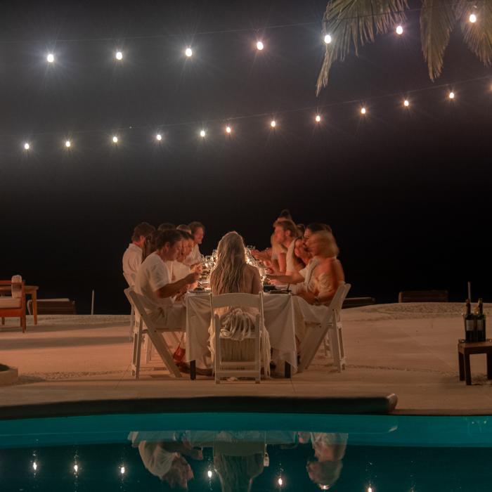 Un groupe de personnes vêtues de blanc dînent à une table sur une plage de sable, la nuit, éclairées par des guirlandes lumineuses, avec une piscine au premier plan qui reflète la scène.
