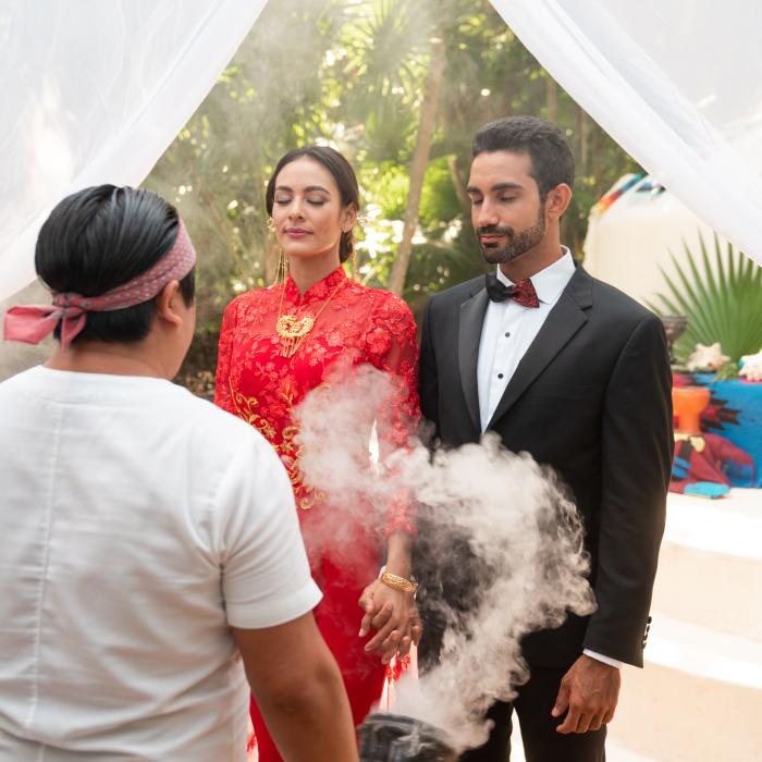 Un marié en smoking et une mariée vêtue d'une robe traditionnelle rouge se tiennent la main lors d'une cérémonie spirituelle accompagnée de fumée.