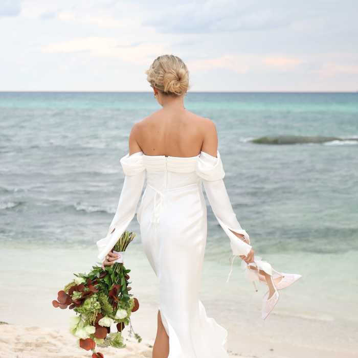 Une mariée en robe blanche, tenant un bouquet et chaussée de talons hauts, sur une plage de sable fin, avec l'océan en arrière-plan.