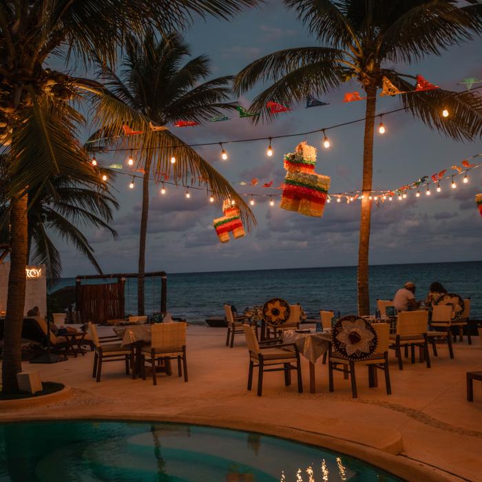À la tombée de la nuit, des palapas, des piñatas et des guirlandes lumineuses décorent un restaurant en bord de mer, avec une piscine au premier plan.