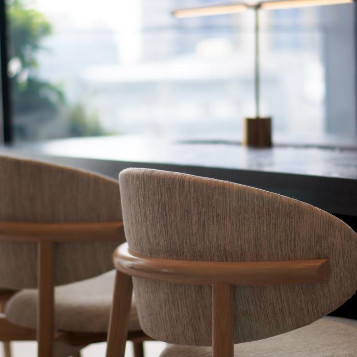 Four upholstered wooden chairs line a dark dining table beside large windows, with a small candle holder on the tabletop.