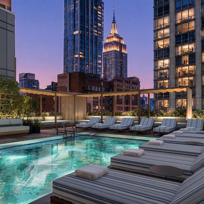 A rooftop pool with striped lounge chairs and planters overlooks illuminated Manhattan skyscrapers at dusk.