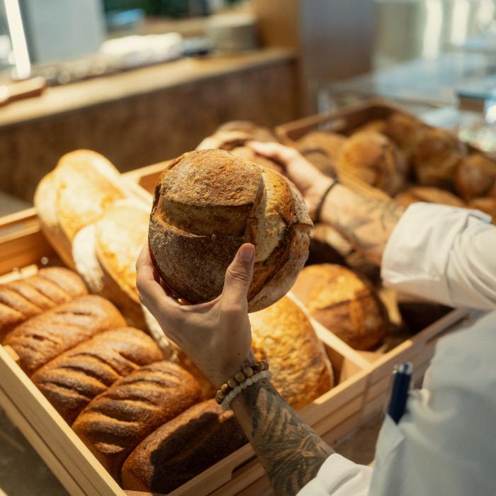 A person in a white shirt holds a round, dark-crusted loaf over wooden bins filled with assorted breads.