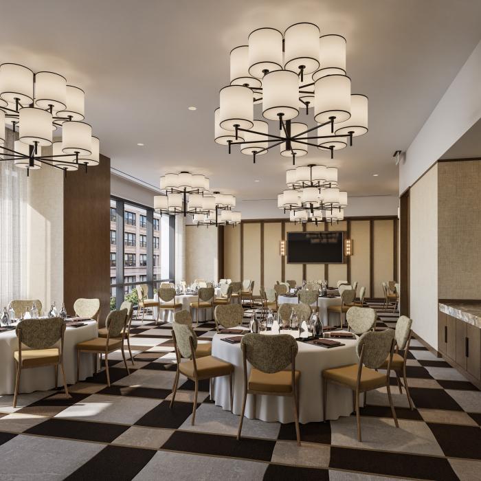 A spacious dining room has round tables set with white cloths and glassware, upholstered chairs, large chandeliers, and tall windows overlooking city buildings.