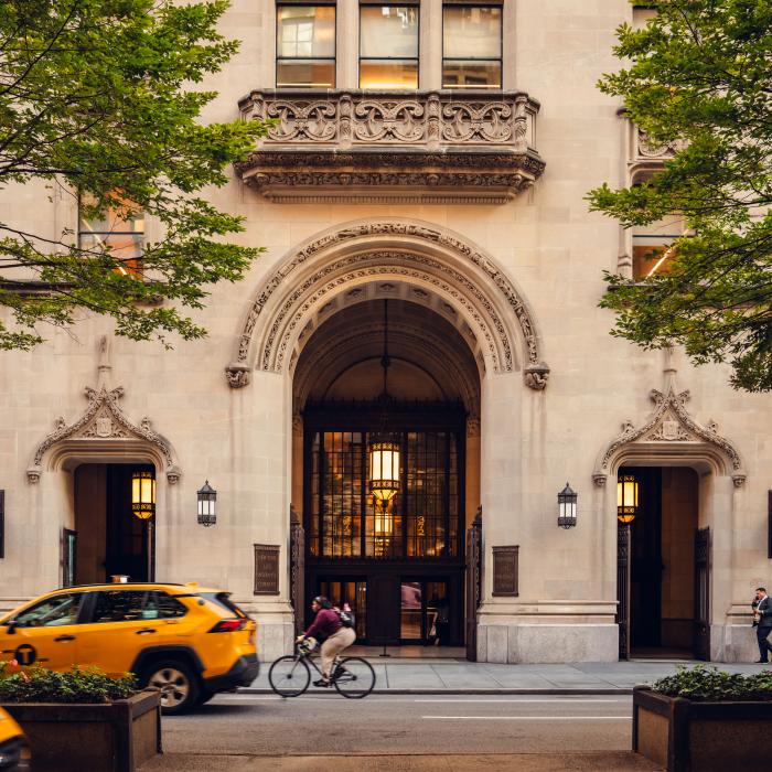 A yellow taxi passes a stone building with an ornate arched entrance as pedestrians walk and a cyclist rides along the street.