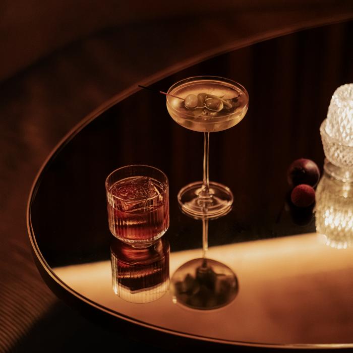 A coupe cocktail garnished with olives and a short glass of amber liquor sit on a round mirrored table beside two dark berries.