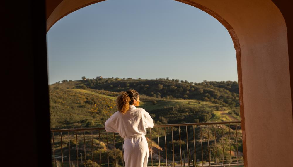 Woman in spa robe looks out over a balcony at rolling green hills.