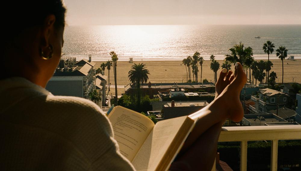 A person sits and reads with their feet up on the railing of their balcony, which overlooks the beach.