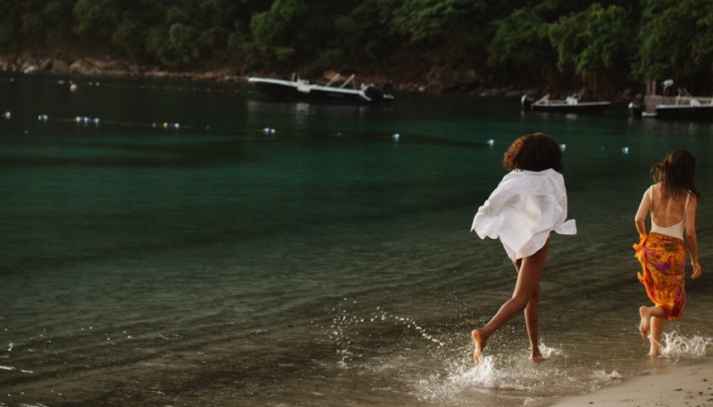 Two women run down a beach, surrounded by the rainforest.