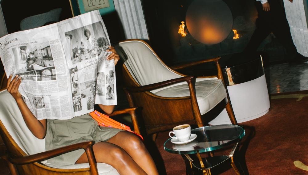 A woman sits reading the newspaper beside a warming fireplace.