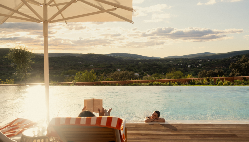 A person sits in a deck chair reading, while their partner lounges in the pool.