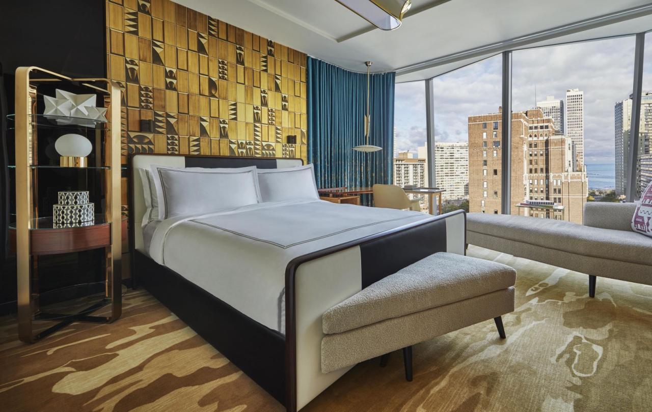 A large bed against an ornate gold accent wall in a Viceroy Chicago guestroom, near a large window overlooking the Gold Coast neighborhood