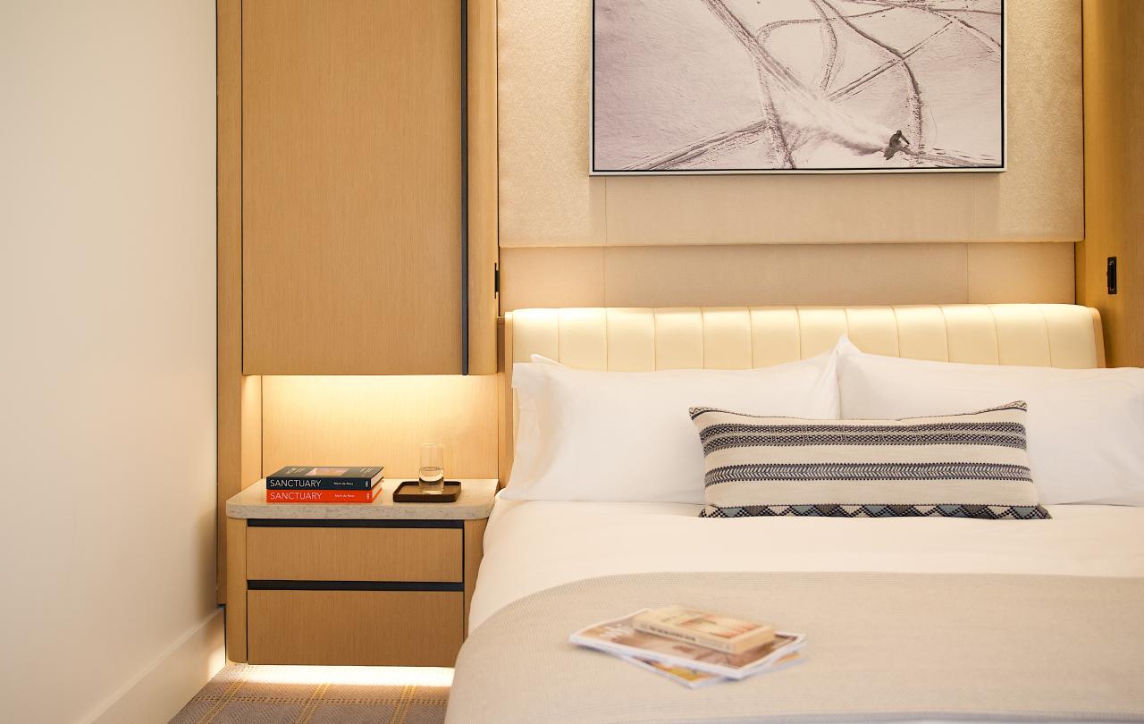 Hotel bed with white linens and a striped throw pillow next to a nightstand with books and water.
