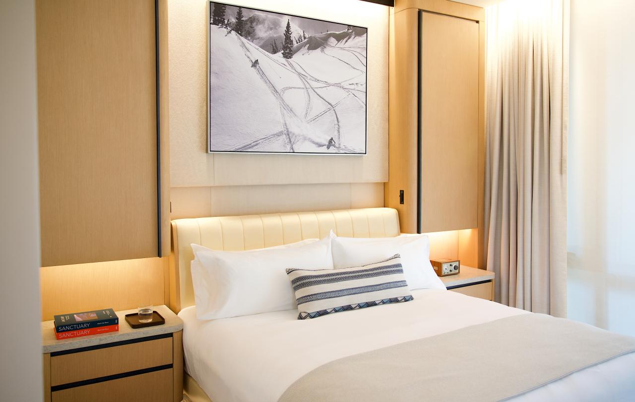 Angle view of a hotel bed with white linens, wooden side tables, and mountain artwork above.