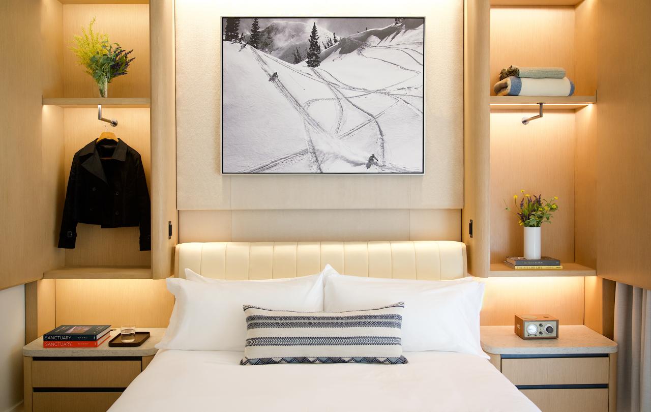 Hotel guest room bed framed by warm wooden built-in shelving with hanging clothing, books, and flowers.