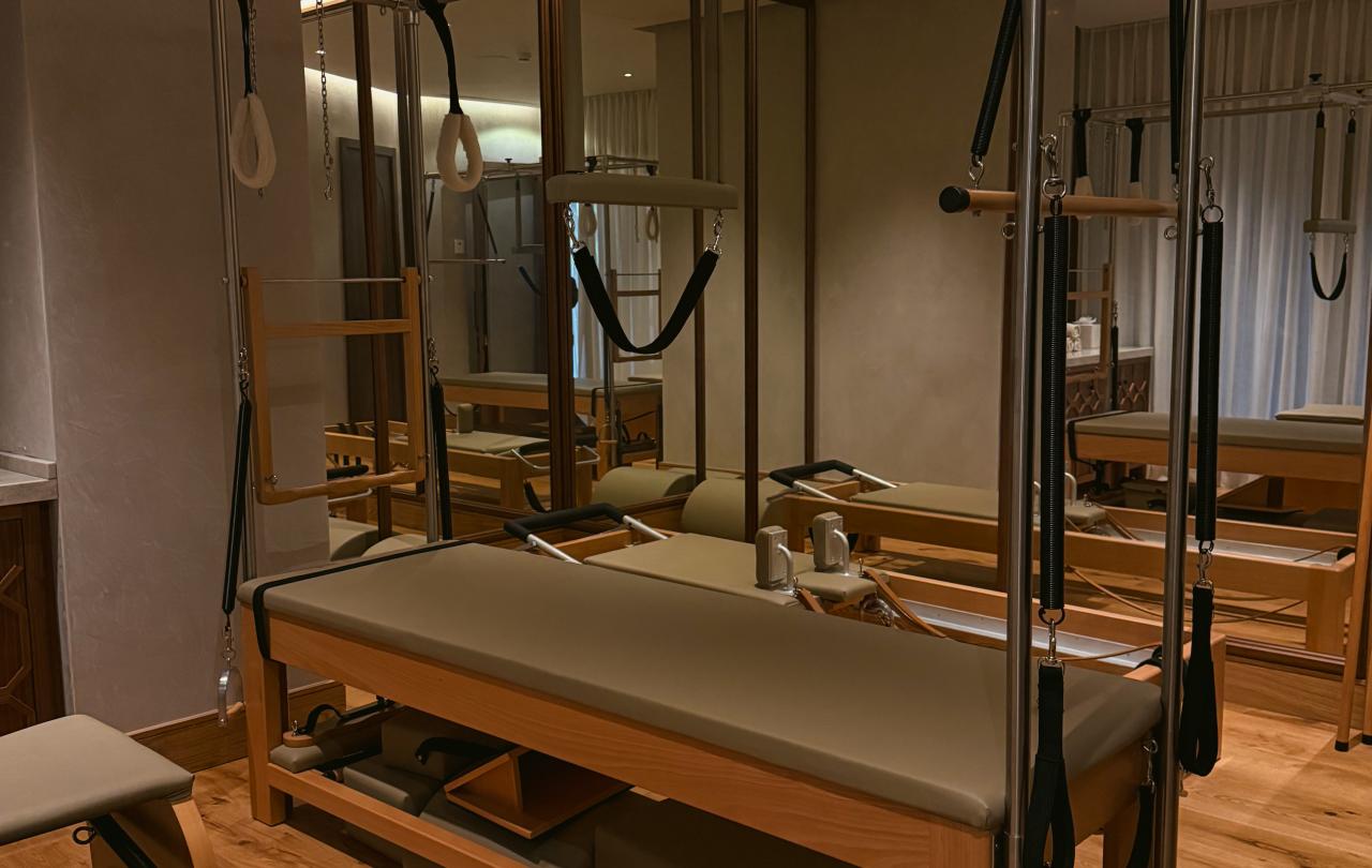 Um aparelho de Pilates com correias e uma superfície almofadada encontra-se num pavimento de madeira, junto a uma parede espelhada.