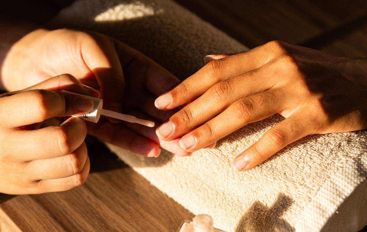 Grande plano de uma manicure a aplicar verniz transparente na mão de uma cliente, apoiada numa toalha.
