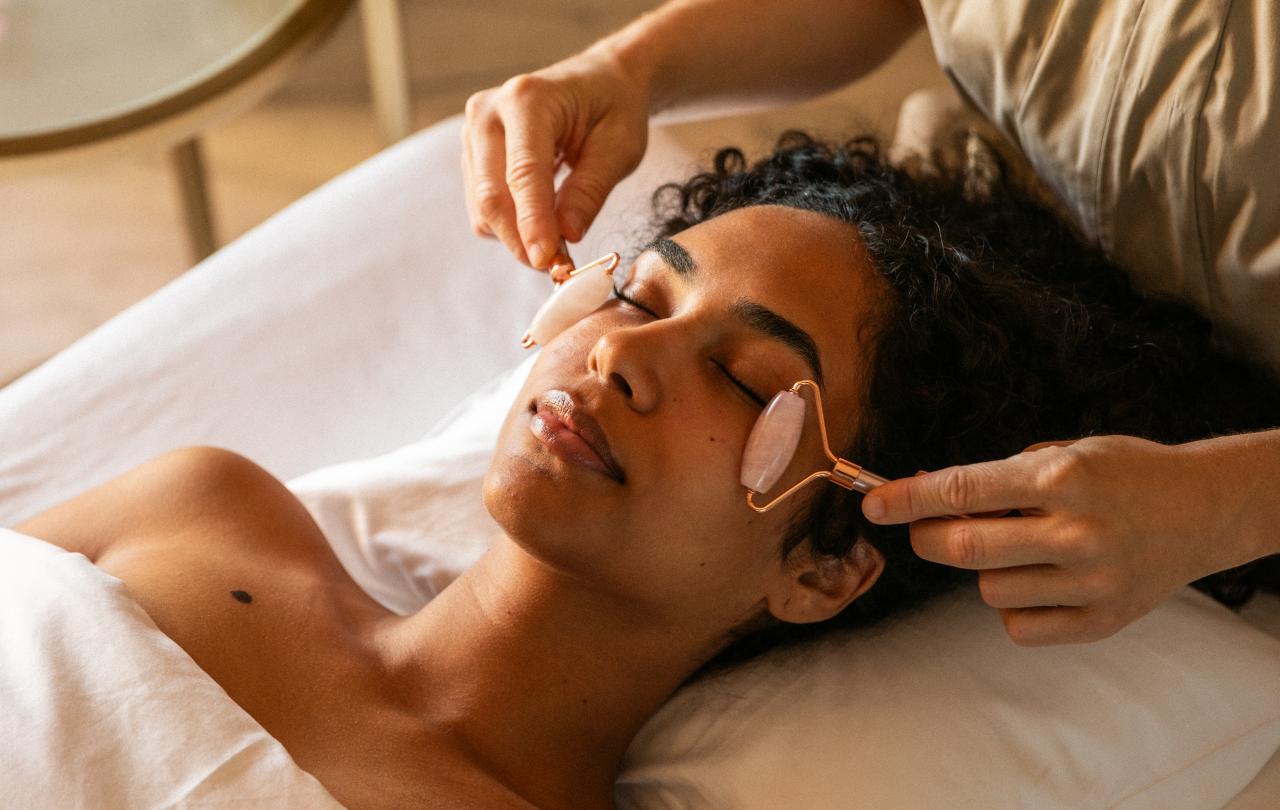 Grande plano de uma esteticista a utilizar rolos de quartzo rosa no rosto relaxado de uma cliente.