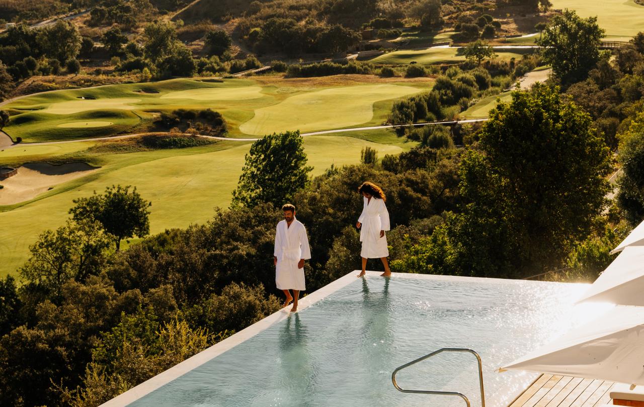 Casal com roupões brancos, de pé à beira de uma piscina infinita com vista para as colinas do campo de golfe.