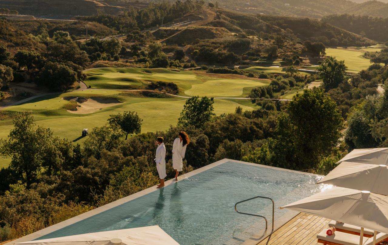 Imagem tirada de um ângulo elevado de uma piscina infinita de um resort e de um campo de golfe, com um casal junto à piscina.
