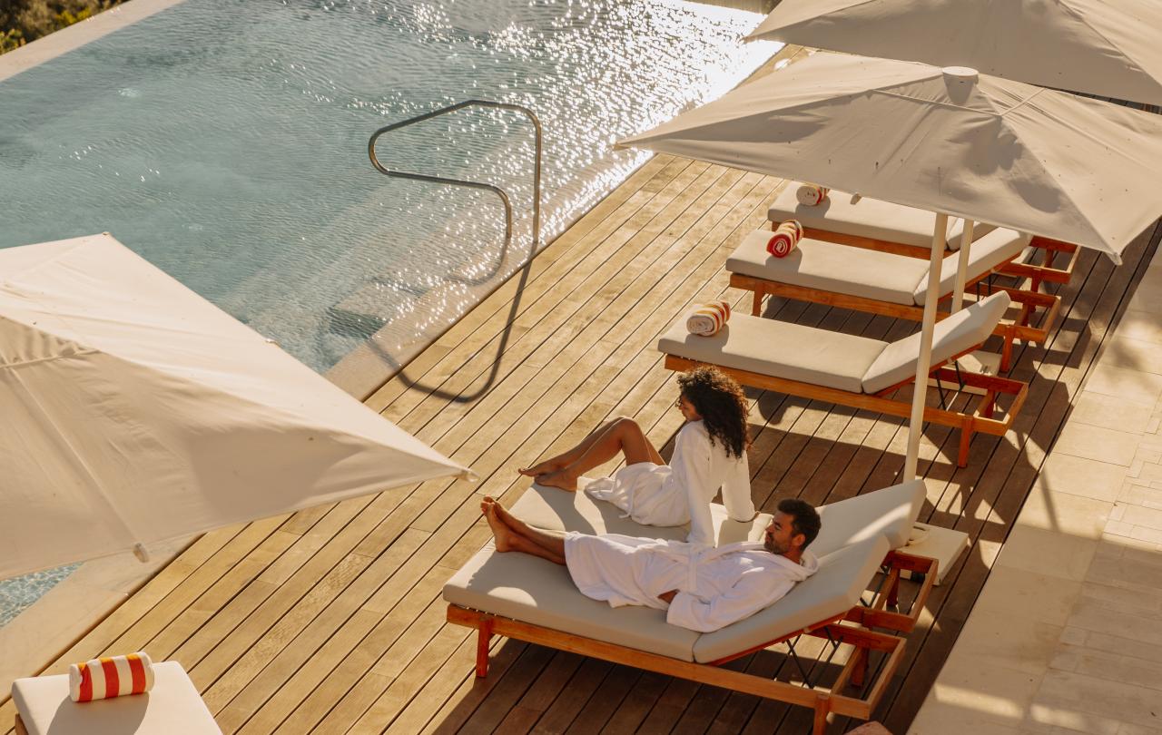 Casal a relaxar junto a uma piscina exterior, num deck de madeira com guarda-sóis brancos.
