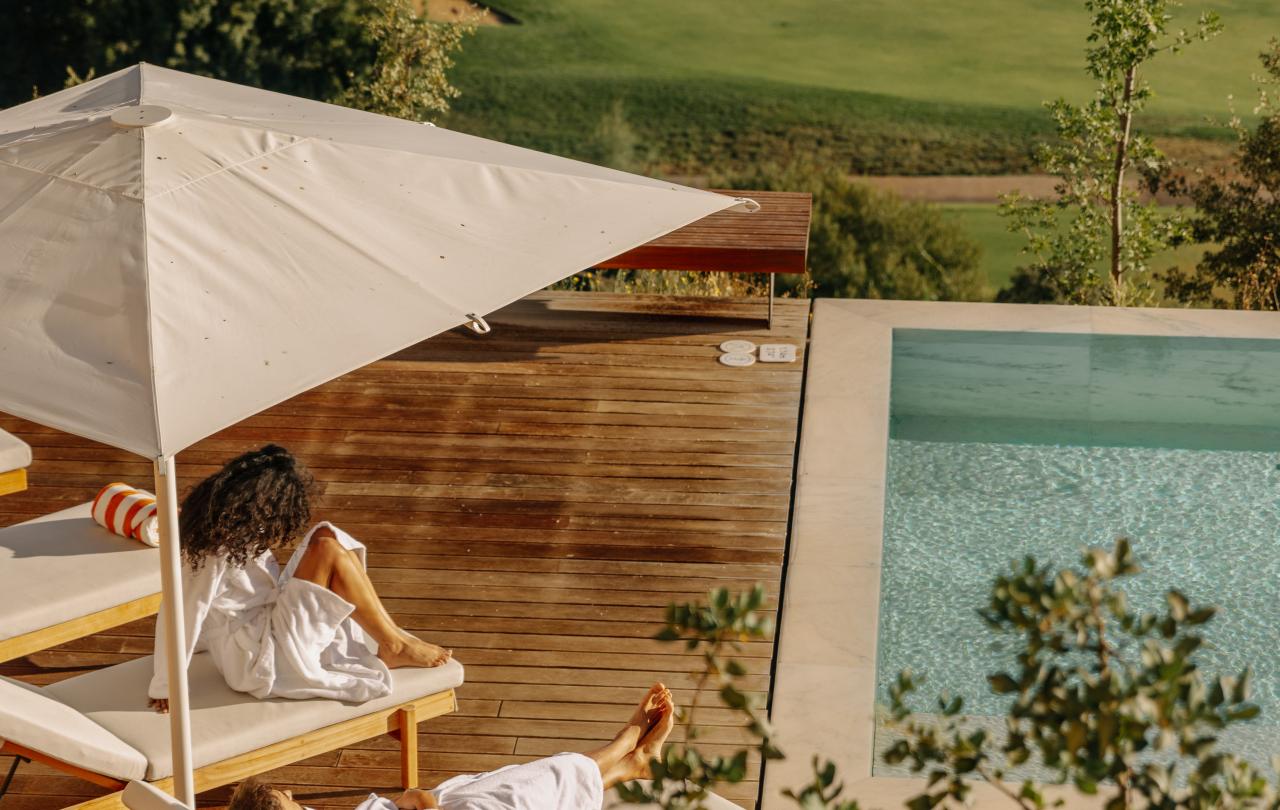 Dois hóspedes, vestidos com luxuosos roupões brancos, repousam em espreguiçadeiras de madeira com vista para um campo de golfe.